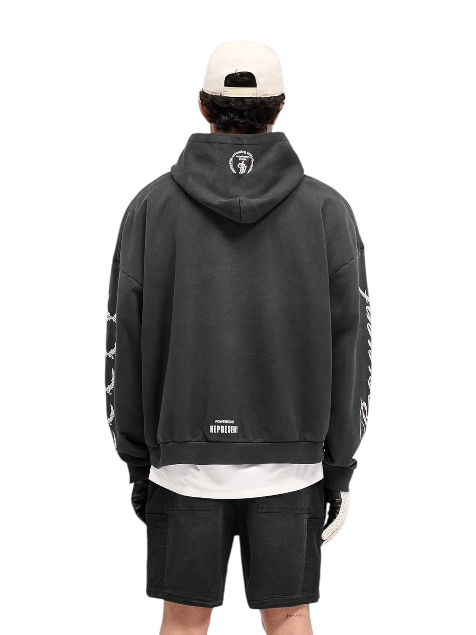 Wearing the Represent MLM41144 World Championship Hoodie in Black by REPRESENT, a person stands against a white background. The hoodie, with graphic prints on the sleeves and back, paired with a baseball cap and black shorts, offers a vintage look through its oversized fit and bold designs.