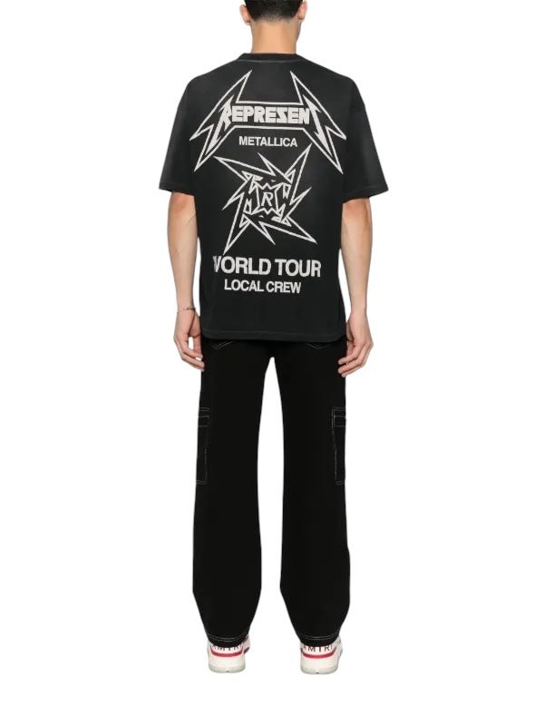 A person stands fashionably on a white background in sleek black pants and a Represent MLM4358 40 Year Anniversary Tee Blk by REPRESENT, featuring "Metallica World Tour Local Crew.