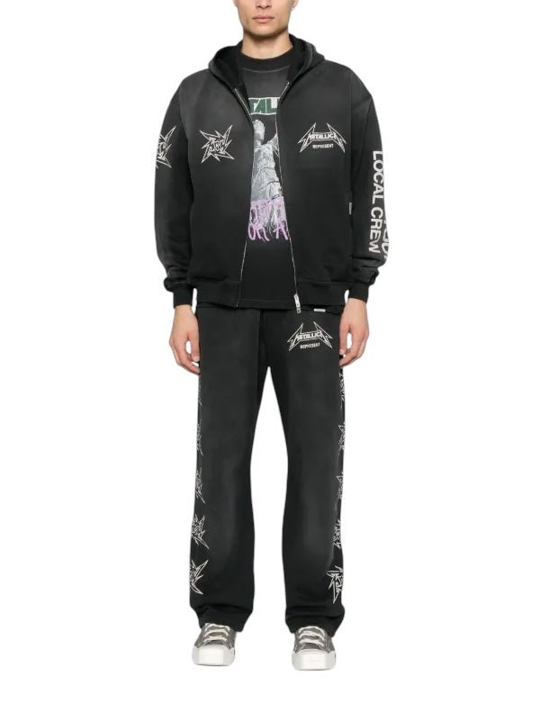 Against a plain white backdrop, the person sports the Represent MLM4370 40 Year Anniversary Zip Hood Blk, featuring iconic Metallica logos. Paired with matching sweatpants and fresh white sneakers, they stand effortlessly cool with hands resting casually by their sides.