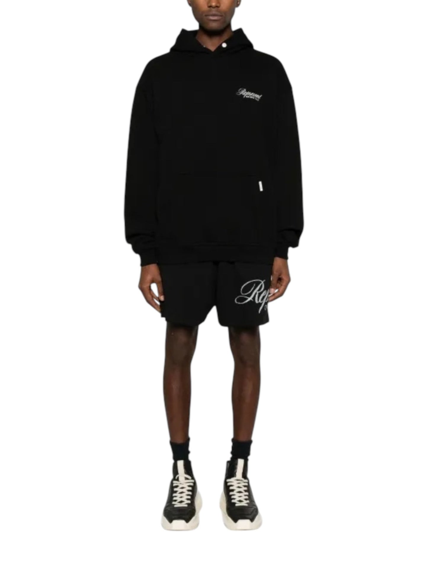 Dressed in a Represent OCM41200 Club Script Hoodie in Black by REPRESENT, along with black shorts and sleek black sneakers featuring white soles, the individual embodies a streetwear vibe against a pristine white backdrop.