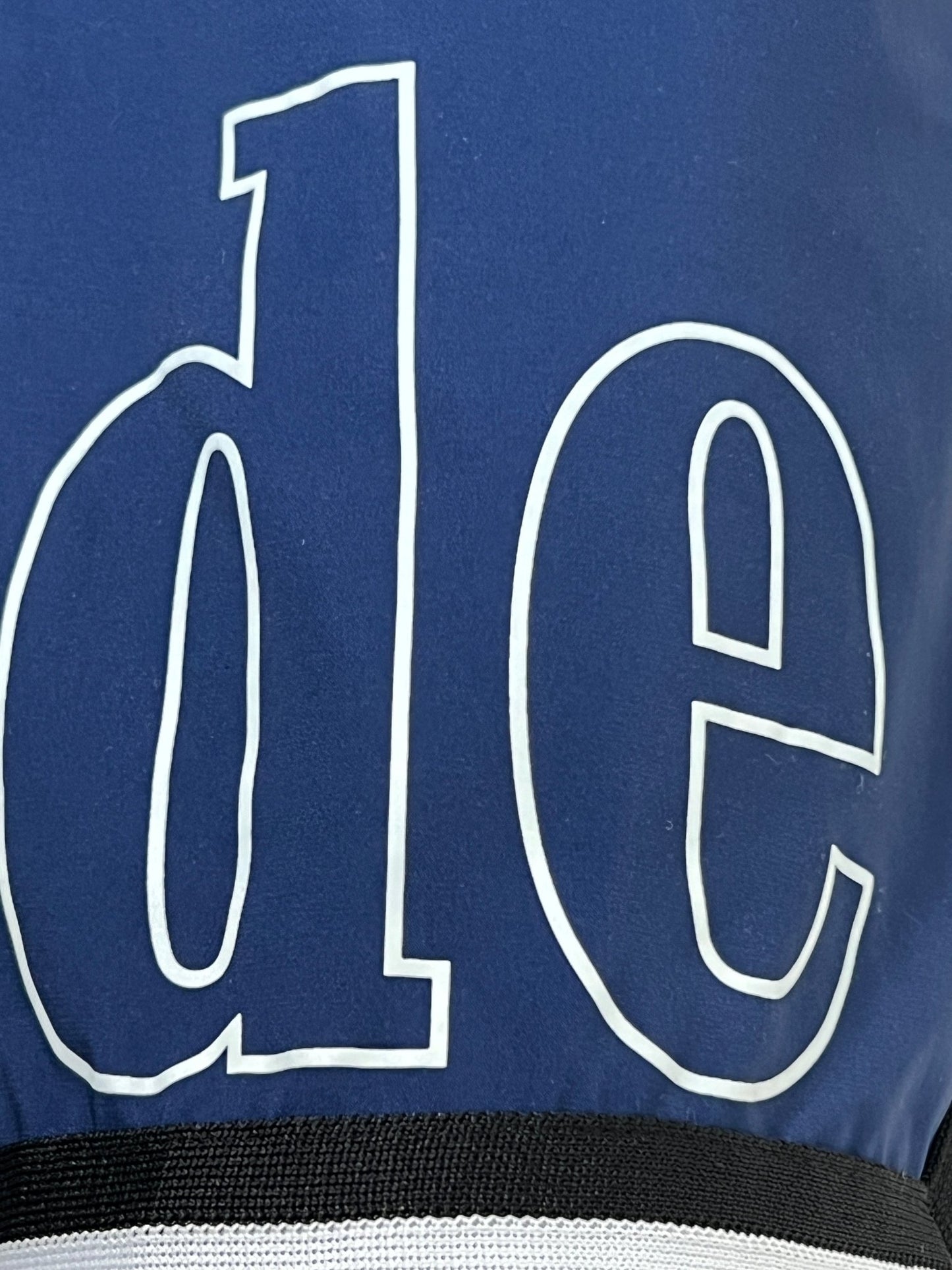 A close-up of bold, white lowercase "d" and "e" in a serif font set against a blue background, featuring a bottom stripe reminiscent of the sleek design found on RHUDE's Rhude Basketball Logo Swim Short Blu.