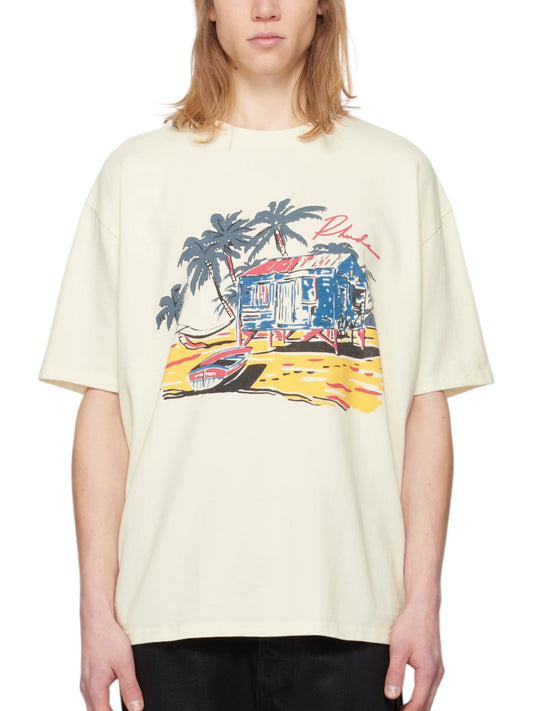 The person wears an RHUDE Beach-Side Tee in white, showcasing a colorful beach house and palm tree design on heavyweight cotton jersey with a comfortable rib-knit crewneck.