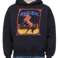 A person wearing the Rhude Cadre Hoodie Black, a black RHUDE cotton fleece hoodie with a bold horse logo and "RHUDE" graphic, paired it effortlessly with blue jeans. The hoodie’s practical kangaroo pocket added both style and functionality to the look.