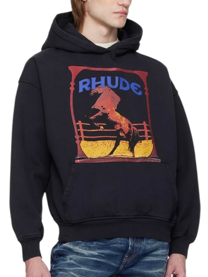 A person wearing a Rhude Cadre Hoodie Black with a horse logo and "RHUDE" graphic, featuring a kangaroo pocket, styled perfectly with blue jeans.