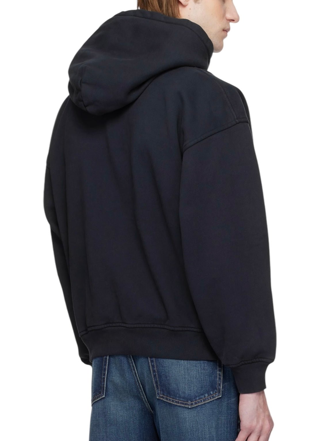 A person wearing the Rhude Cadre Hoodie Black, a sleek black cotton fleece piece by RHUDE featuring a subtle logo graphic, pairs it with blue jeans and faces partially away from the camera.