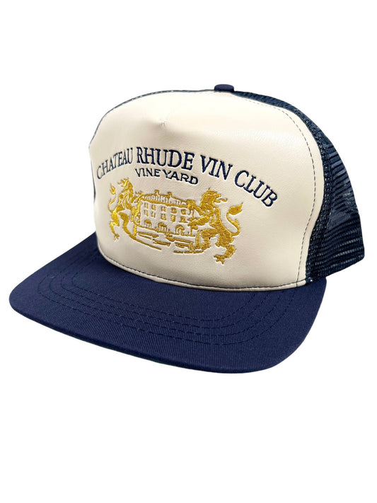 The RHUDE CELLIER TRUCKER HAT NAVY/CREAM features a black mesh back and a navy blue brim. The cream-colored front panel displays a "Chateau Rhude Vin Club Vineyard" emblem, accentuated by an embroidered leather detail with a gold design of a chateau and lions.