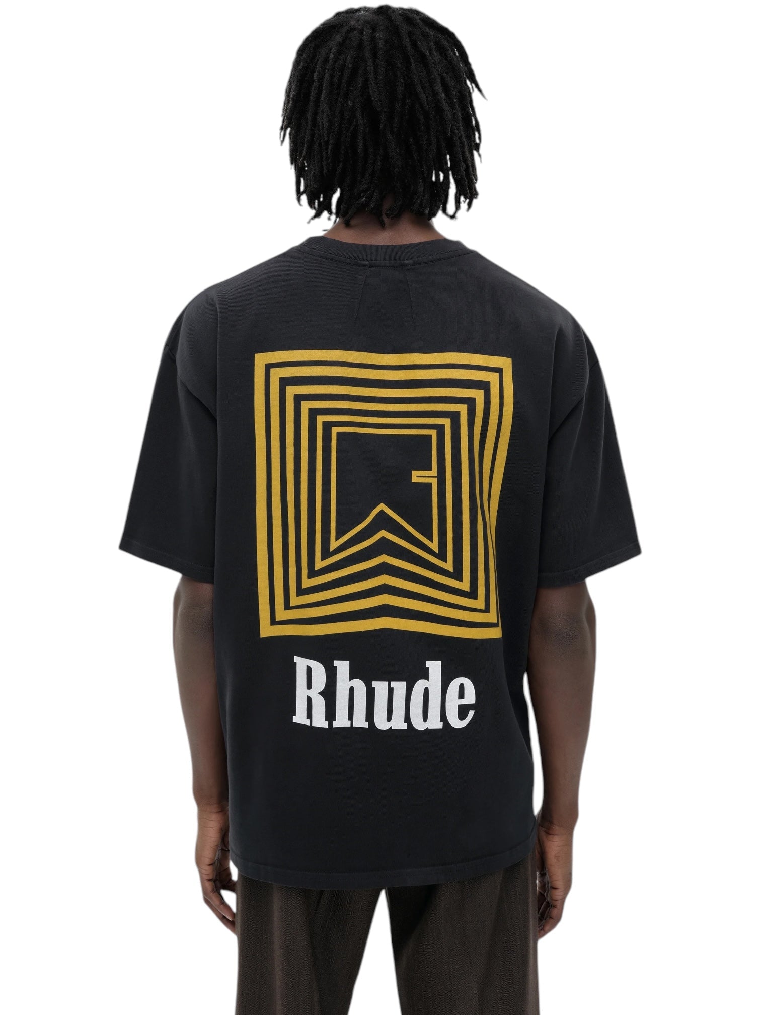 A person is wearing the Rhude Chevron Logo Repeat Tee in vintage black, featuring a geometric yellow design and "Rhude" text on the back, along with the striking Rhude Chevron Box logo.