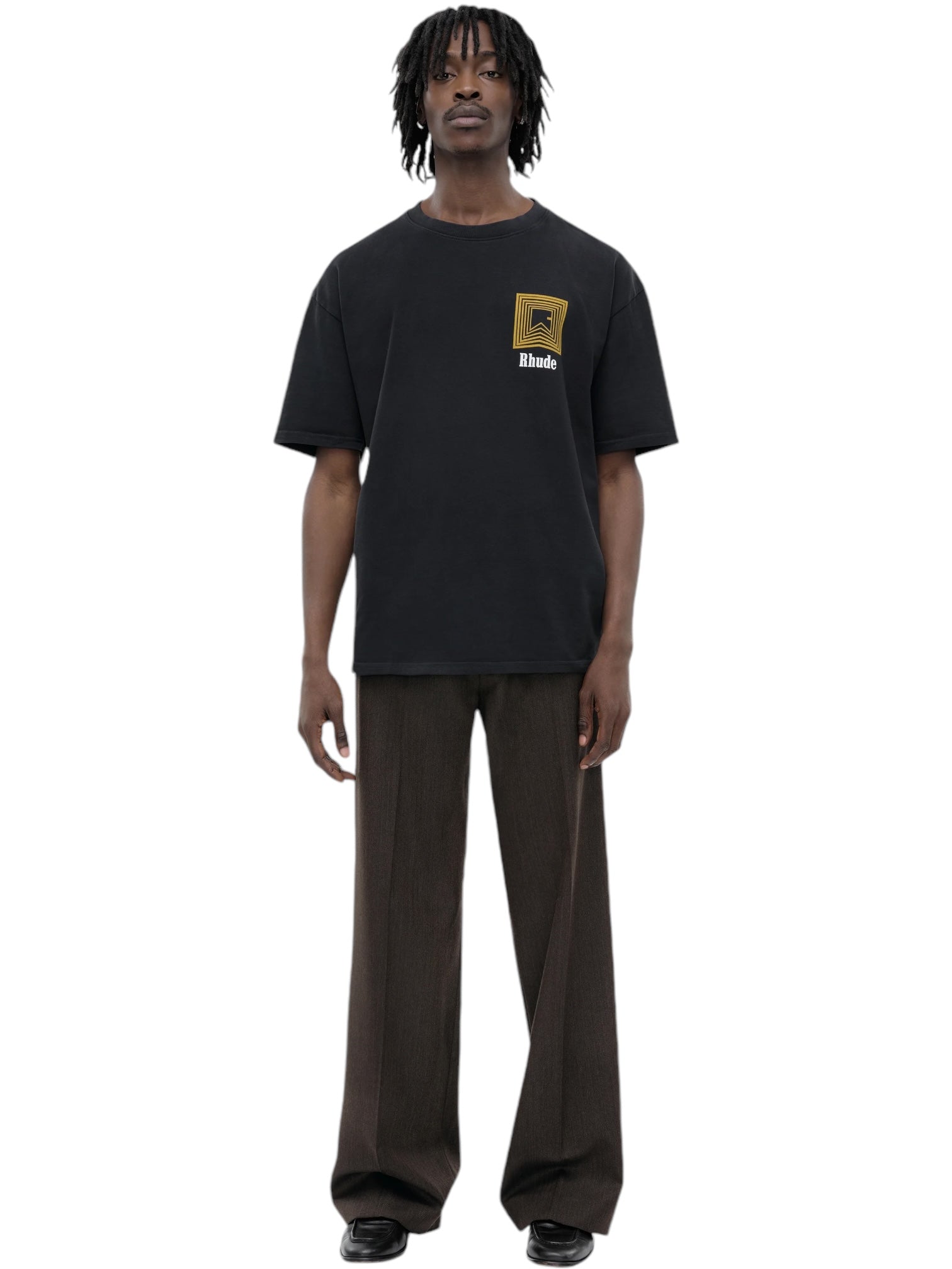 A person stands on a plain white background, wearing the Rhude Chevron Logo Repeat Tee in vintage black from RHUDE, paired with vintage wash brown wide-leg pants and black shoes.