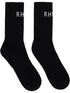 The Rhude Classic Logo Socks by RHUDE are black, calf-high, and ribbed with a white "RH" jacquard logo near the top.