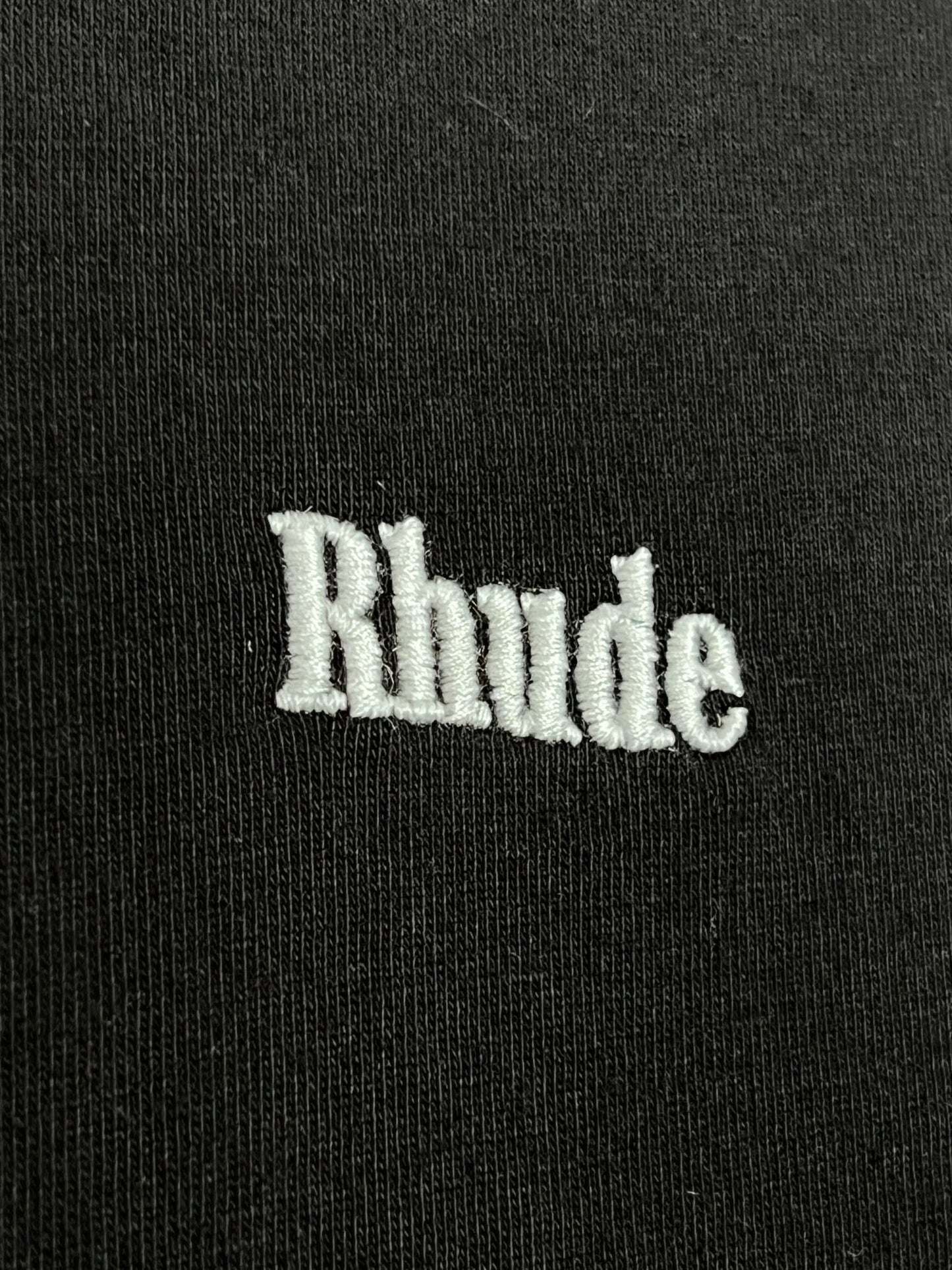 A close-up of the Rhude Classique Reverse Ls Tee Black by RHUDE, showcasing 100% cotton fabric with "Rhude" intricately embroidered in white thread, encapsulating the essence of this long sleeve t-shirt.