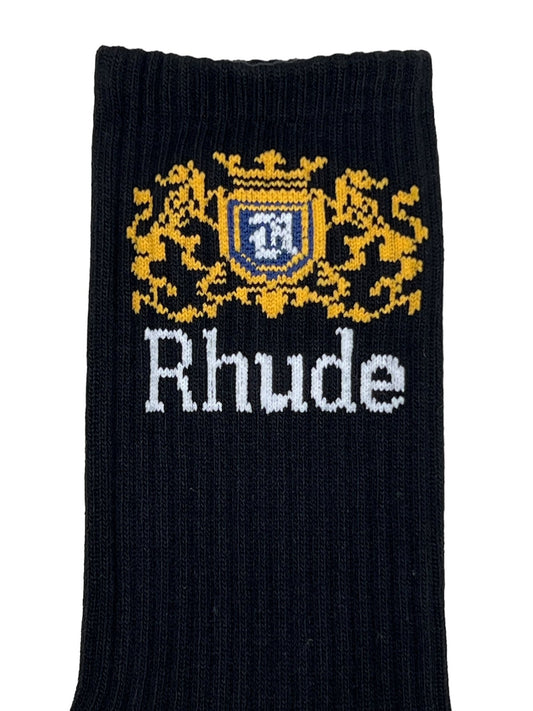 Close-up of black fabric with "Rhude" written in white text, below a golden emblem featuring detailed gold foliage and a blue shield. This intricate jacquard logo design is part of the RHUDE RHUDE CREST LOGO SOCK BLK WHITE YELLOW collection.