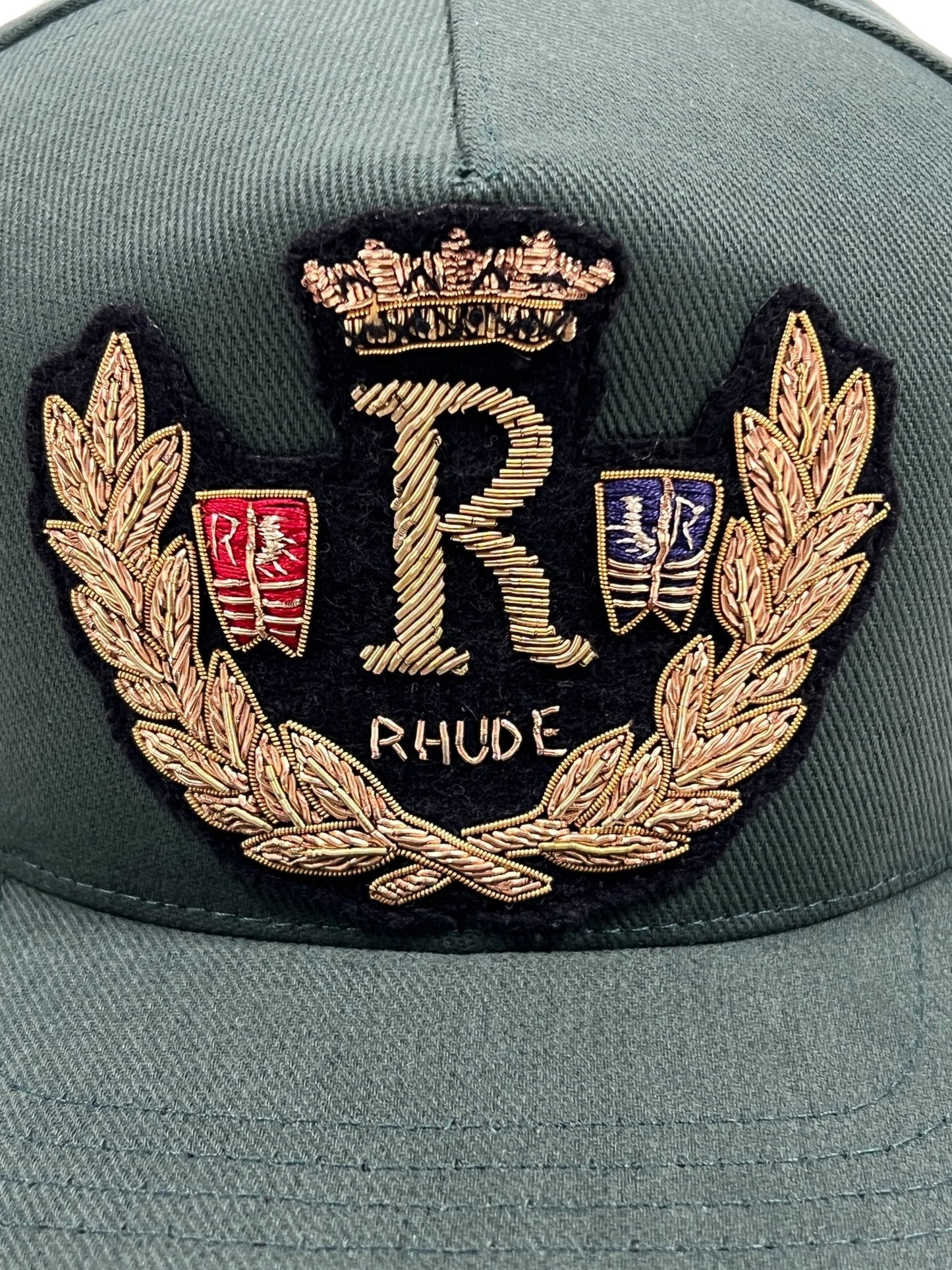 The RHUDE DIANA CREST HAT in green features an embroidered emblem showcasing a crown, a large "R," a laurel wreath, and the word "RHUDE." It also includes small red and blue crests on either side of the "R.