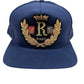 The RHUDE DIANA CREST HAT NAVY by RHUDE is a navy blue baseball cap made from 100% cotton, adorned with a gold embroidered "R" accompanied by a crown and laurel wreath, plus red and blue shield accents.