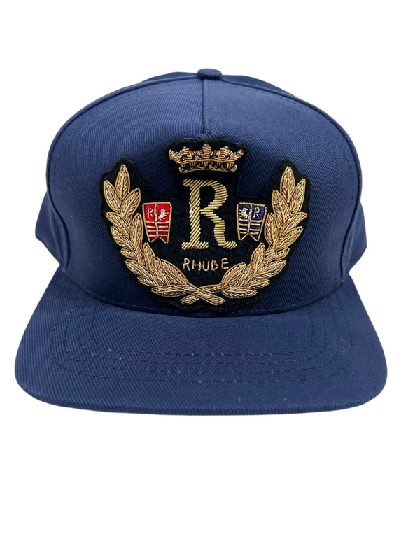 The RHUDE DIANA CREST HAT NAVY by RHUDE is a navy blue baseball cap made from 100% cotton, adorned with a gold embroidered "R" accompanied by a crown and laurel wreath, plus red and blue shield accents.