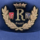 Close-up of the RHUDE DIANA CREST HAT in navy, featuring an embroidered design with the letter "R," gold laurel leaves, a crown, and the word "RHUDE." Two small shields with heraldic symbols flank the central "R." Made from 100% cotton, this cap showcases a crest-like elegance perfect for any stylish collection.
