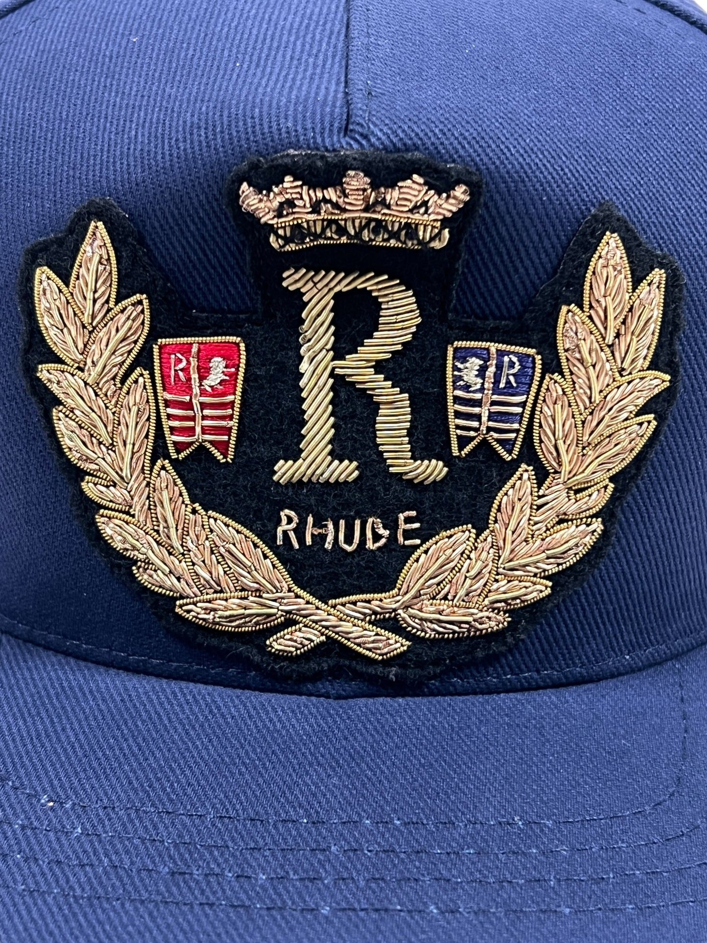 Close-up of the RHUDE DIANA CREST HAT in navy, featuring an embroidered design with the letter "R," gold laurel leaves, a crown, and the word "RHUDE." Two small shields with heraldic symbols flank the central "R." Made from 100% cotton, this cap showcases a crest-like elegance perfect for any stylish collection.