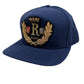 The RHUDE DIANA CREST HAT in navy blue is made from 100% cotton and showcases a gold embroidered design with the letter "R" at the center and "Rhude" beneath it.