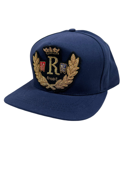 The RHUDE DIANA CREST HAT in navy blue is made from 100% cotton and showcases a gold embroidered design with the letter "R" at the center and "Rhude" beneath it.