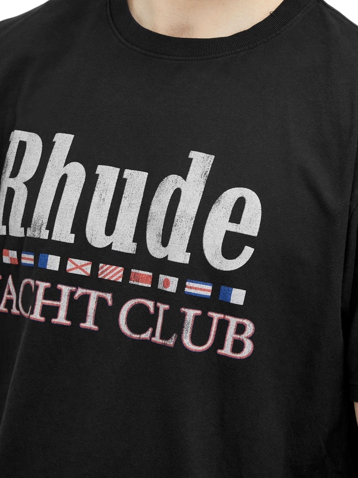 A person wearing the RHUDE FLAG TEE BLK, a black graphic tee by RHUDE with "Rhude Yacht Club" and nautical flags on the front.