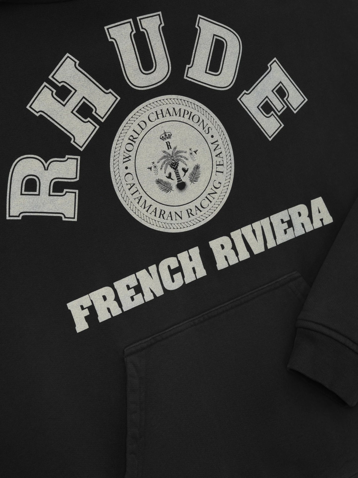 The Rhude French Riviera Hood Black by RHUDE features a kangaroo front pocket and is styled with white "RHUDE" text, a "World Champions Catamaran Racing Team" emblem, and a "FRENCH RIVIERA" design.