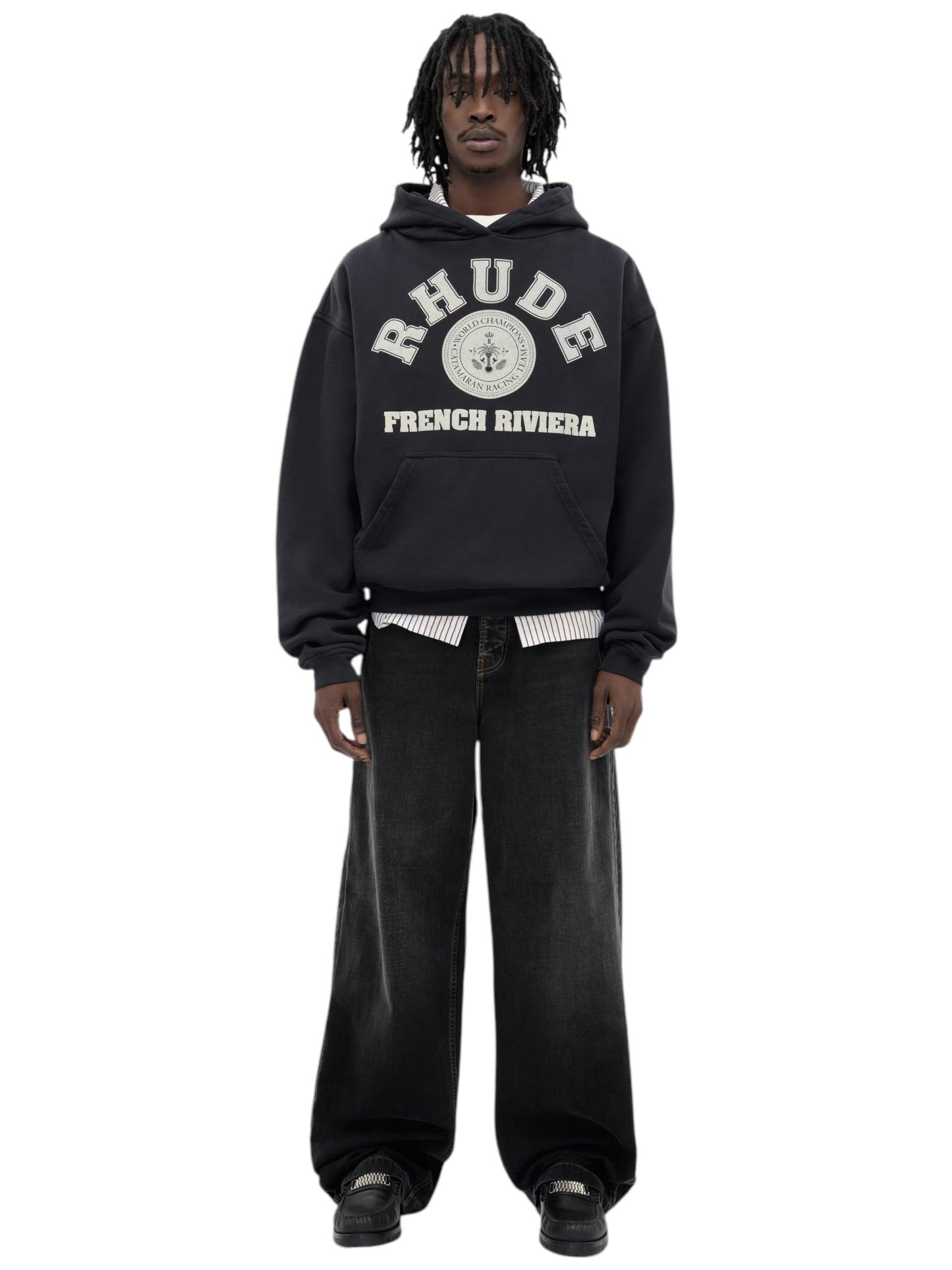 A person wearing a RHUDE "Rhude French Riviera Hood Black" hoodie with a kangaroo pocket, layered shirts underneath, and black wide-leg pants.