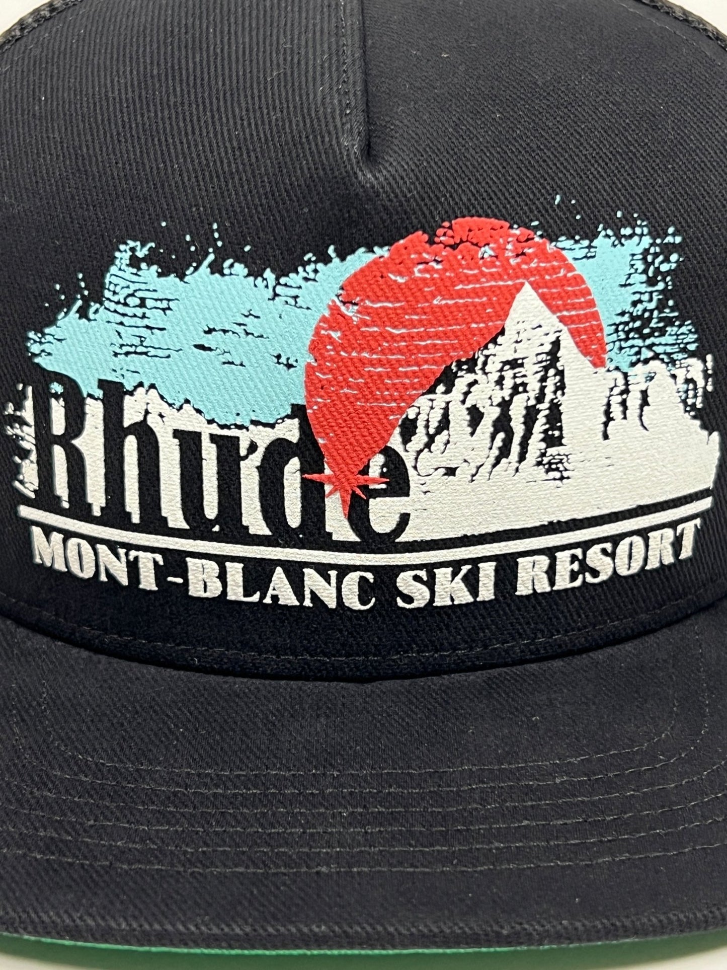 A black RHUDE MONT-BLANC TRUCKER HAT BLACK with an adjustable snapback showcases a printed design featuring the words "Rhude Mont-Blanc Ski Resort", a mountain silhouette, and a red sun.