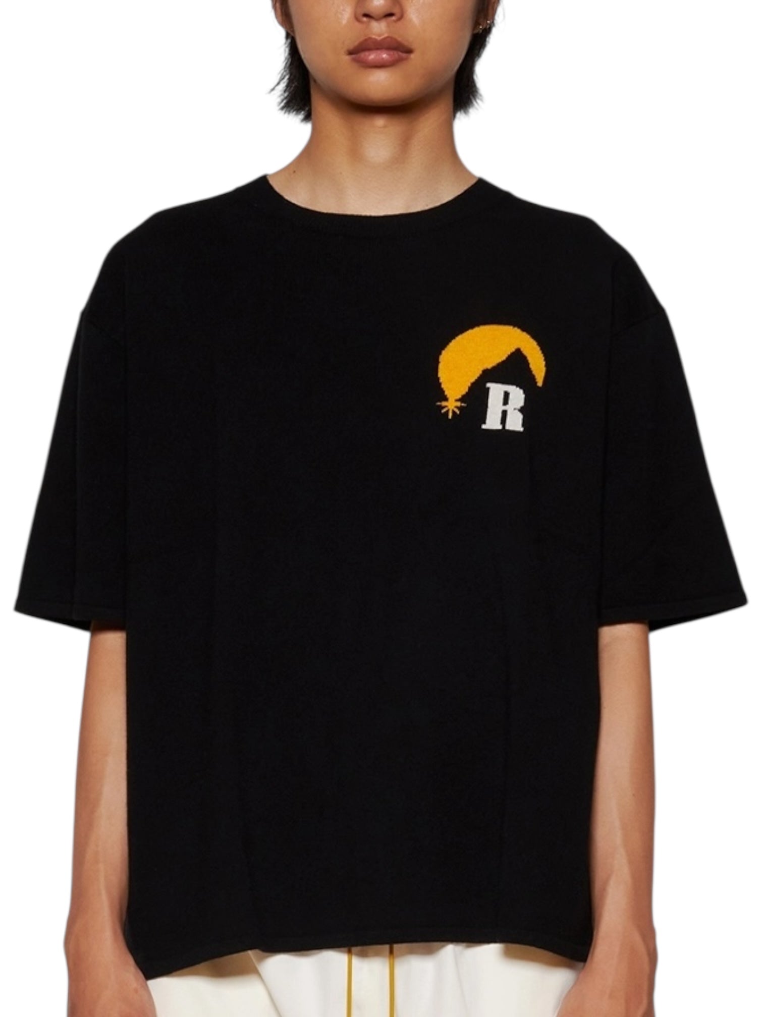 A person wears the RHUDE Moonlight Knit Tee Black/Cream from Fall/Winter 2024, featuring a yellow crescent and white "R" logo on the left chest, standing against a plain white background.