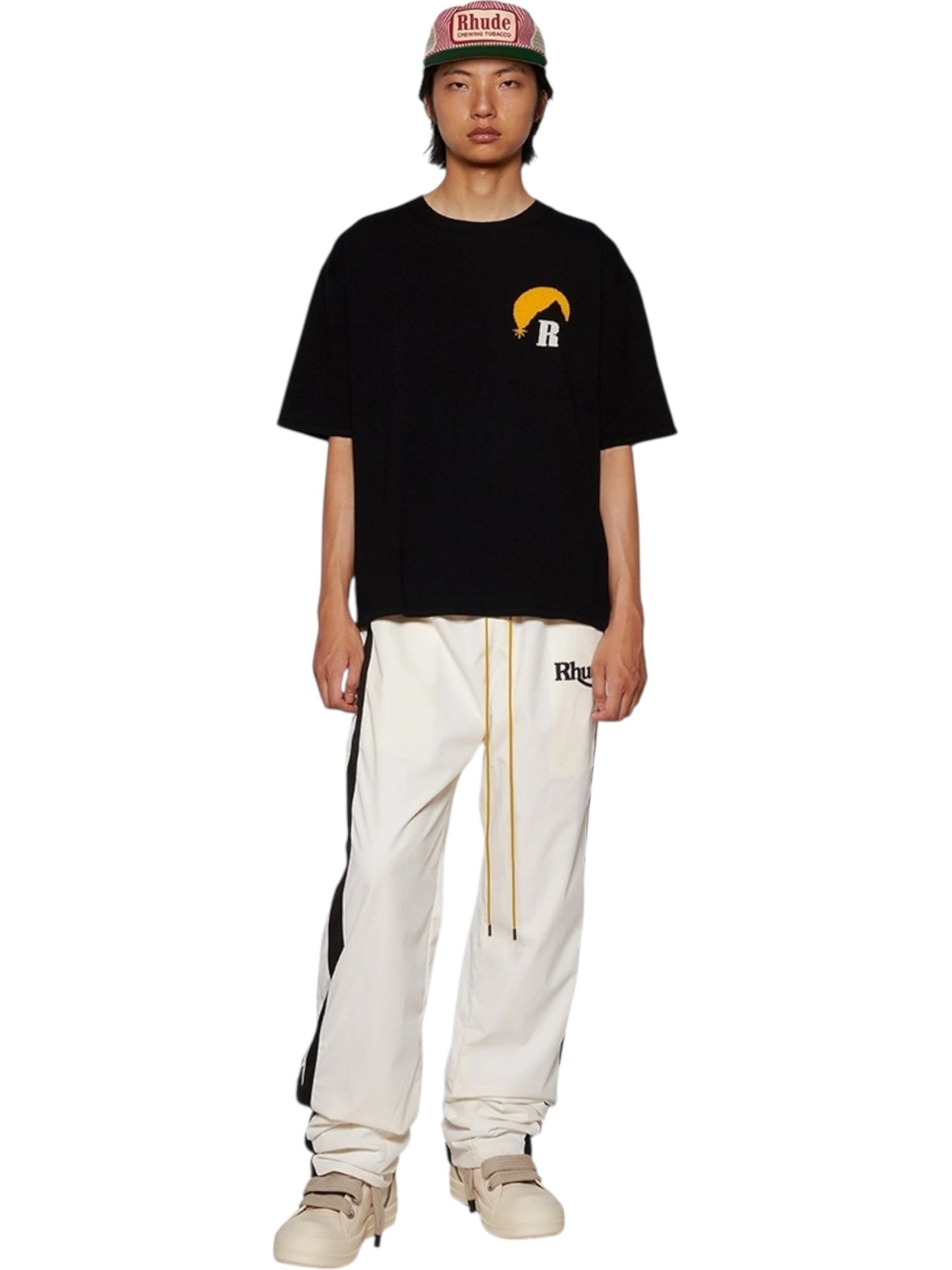 A person stands against a white background wearing a RHUDE Moonlight Knit Tee Black/Cream from Fall/Winter 2024, paired with white track pants featuring black stripes, cream sneakers, and a red RHUDE cap.