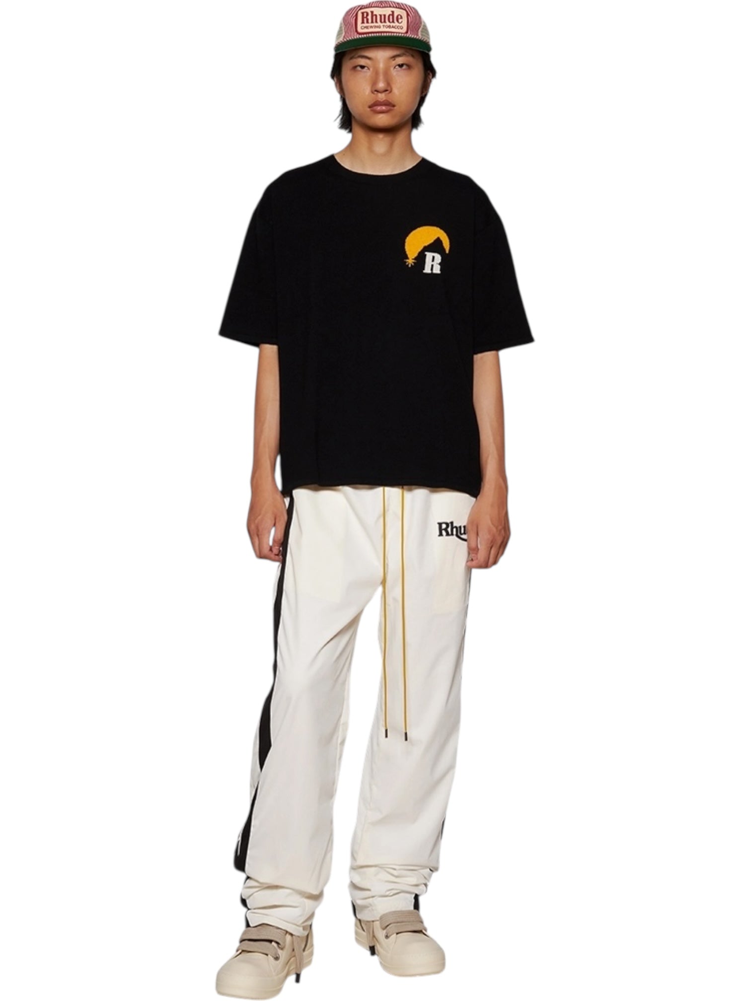 A person stands against a white background wearing a RHUDE Moonlight Knit Tee Black/Cream from Fall/Winter 2024, paired with white track pants featuring black stripes, cream sneakers, and a red RHUDE cap.