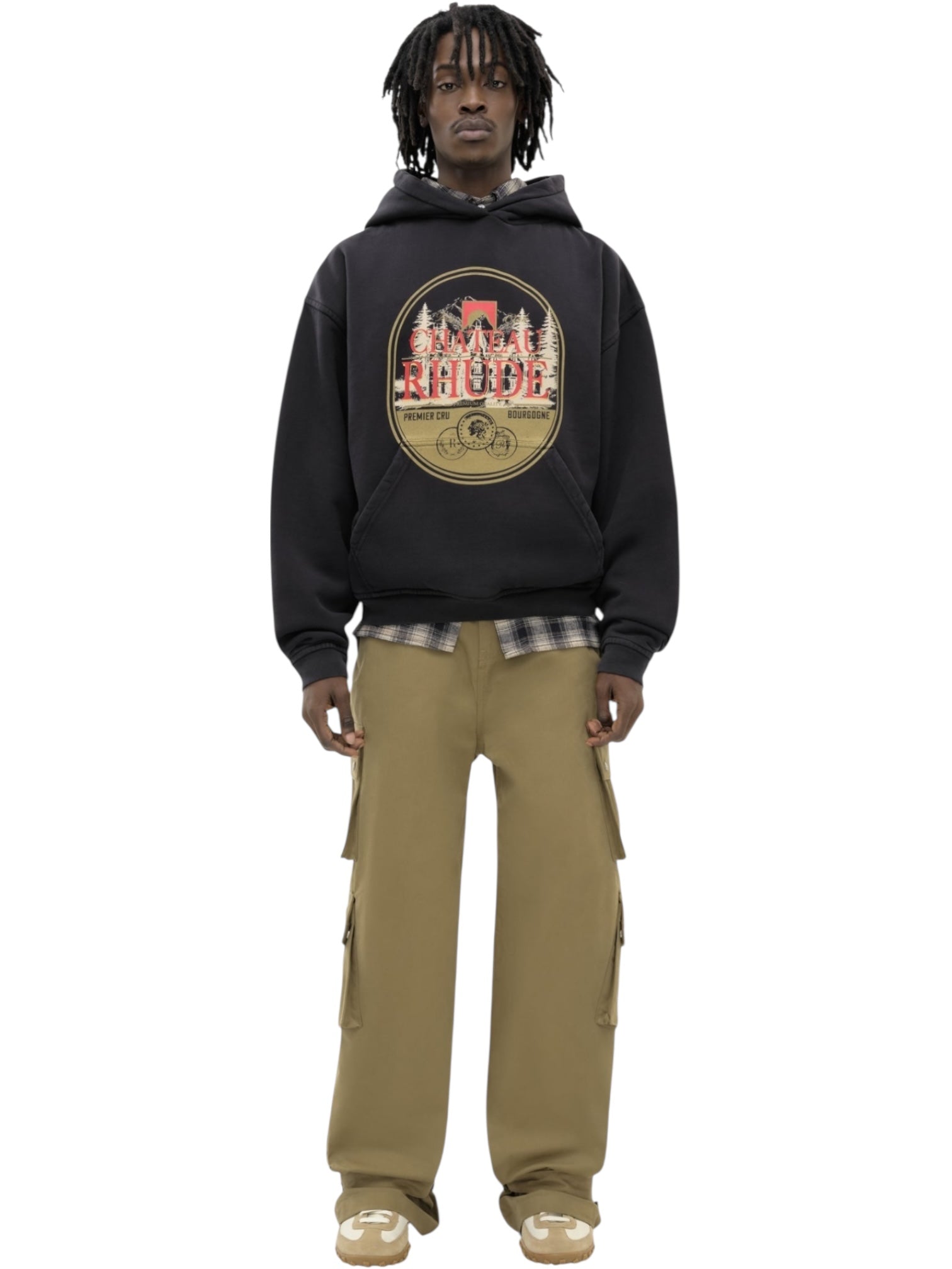A man faces forward wearing the RHUDE Rhude Premier Hoodie Vtg Black with a kangaroo pocket and printed graphic, a plaid shirt underneath, khaki cargo pants, and beige sneakers.