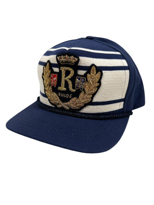 The RHUDE ROPE CREST STRUCTURE HAT NAVY by RHUDE is a navy blue and white striped hat showcasing an embroidered "Rhude" logo, elegantly adorned with a gold laurel wreath and shield crest on the front.