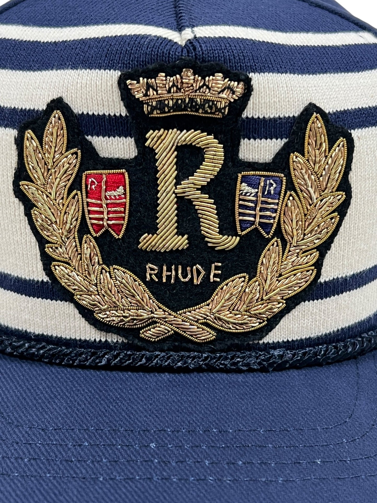 Close-up of a navy blue and white striped RHUDE ROPE CREST STRUCTURE HAT NAVY, featuring an embroidered emblem. The crest is adorned with a gold crown, a prominent gold "R," red and blue shields, laurel leaves, and the word "RHUDE" beneath.