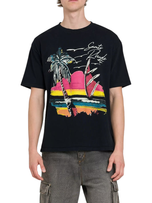 A person is wearing a black Rhude Saint-Rhude Tee by RHUDE, made from cotton jersey, featuring a colorful sunset graphic with palm trees and a sailboat, paired with gray cargo pants.