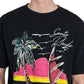 A person wearing a black Rhude Saint-Rhude Tee from RHUDE, made of soft cotton jersey and featuring a vibrant graphic print of a palm tree, waves, and the text "Santo Dhude.