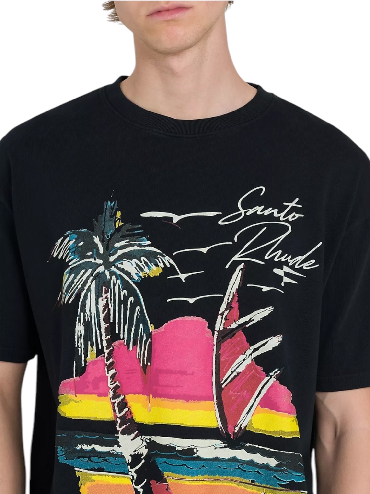 A person wearing a black Rhude Saint-Rhude Tee from RHUDE, made of soft cotton jersey and featuring a vibrant graphic print of a palm tree, waves, and the text "Santo Dhude.