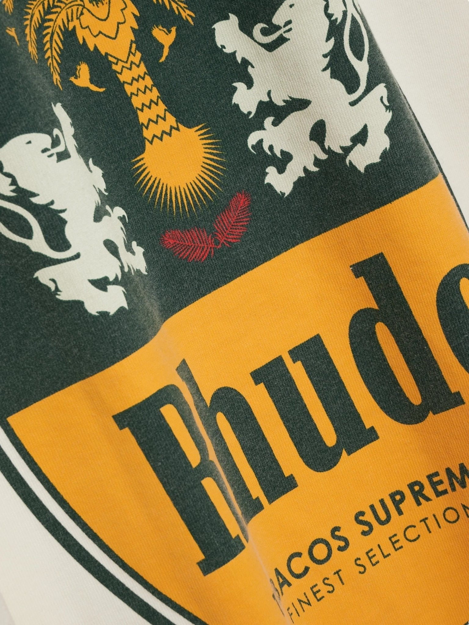 Close-up of RHUDE Tabaco Tee Vtg White showing a crest with two white lions, a yellow palm tree, and "Rhude" in bold on a yellow background printed on vintage wash cotton fabric.