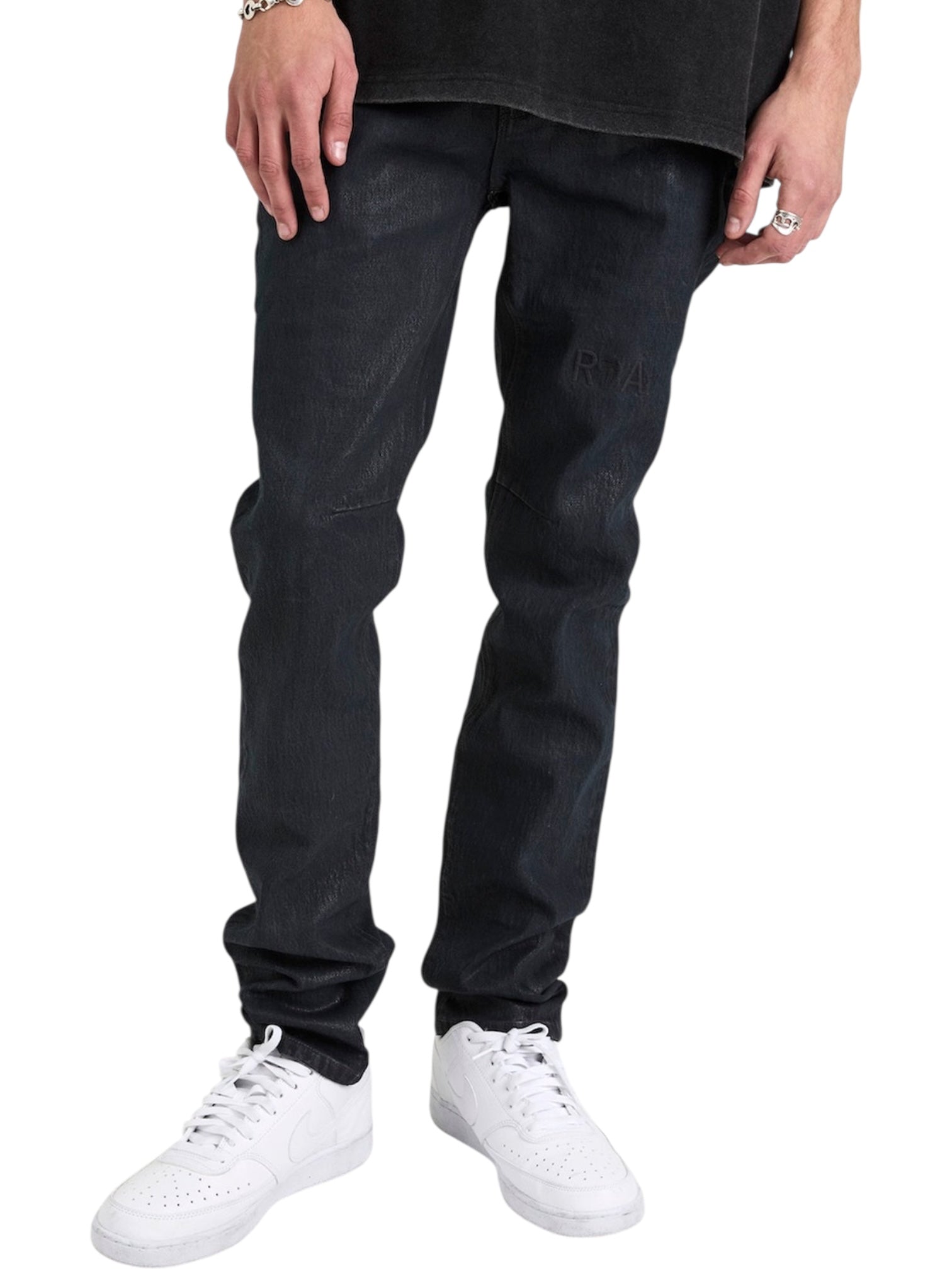 Someone wearing RTA Clayton Coated Black jeans from RTA, a black shirt, white sneakers, a bracelet, and rings stands against a white background.