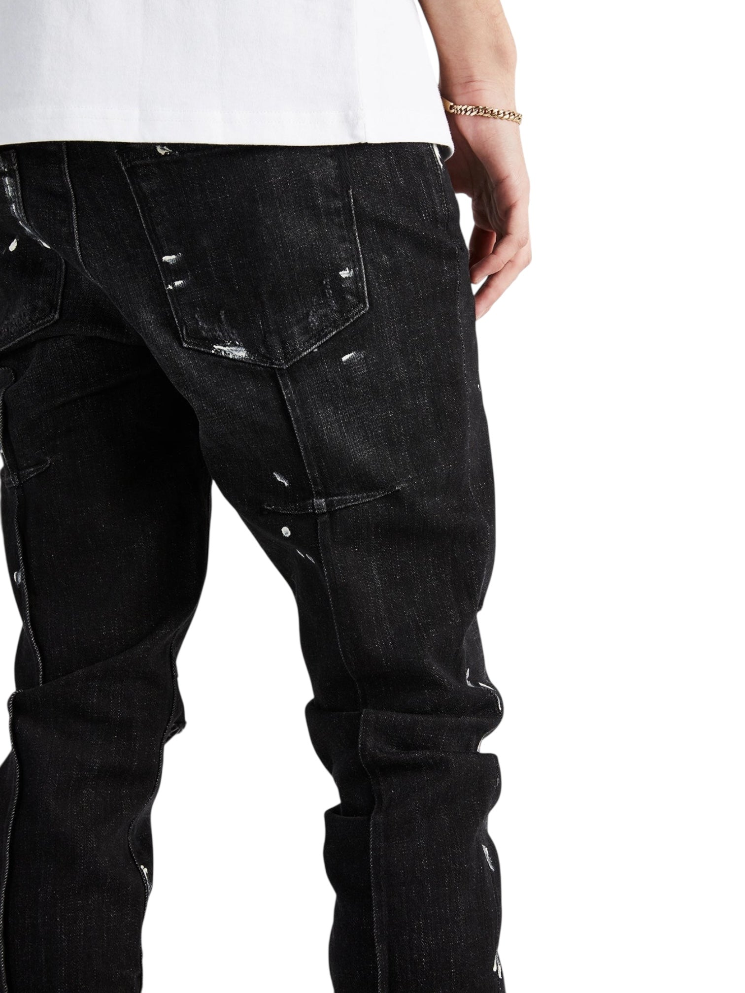 Model wearing RTA Clayton Jean Distressed Charcoal Paint Black, a skinny 5-pocket jean with white paint splatter from RTA; styled with a white shirt, showing only the lower torso and upper legs from the back.