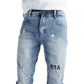 A person wearing RTA Clayton Jean Distressed White Paint Blue by RTA, featuring light blue distressed denim with "RTA" printed on the left thigh, paired with a white t-shirt tucked in at the waist.