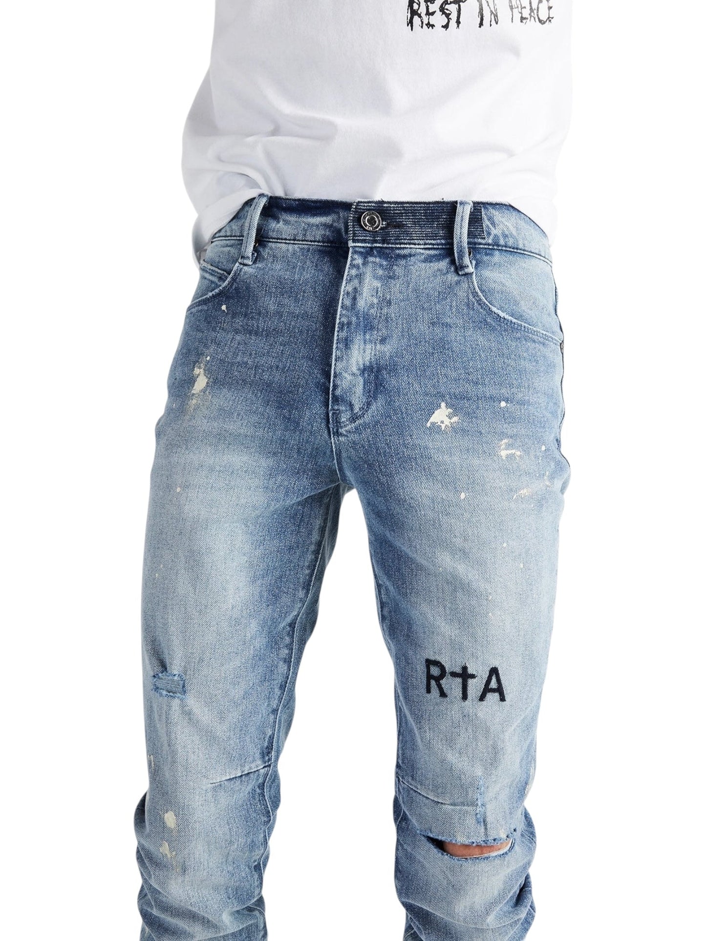 A person wearing RTA Clayton Jean Distressed White Paint Blue by RTA, featuring light blue distressed denim with "RTA" printed on the left thigh, paired with a white t-shirt tucked in at the waist.