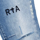 Close-up of RTA Clayton Jean Distressed White Paint Blue, featuring distressed white denim with blue paint accents and a black "R†A" logo embroidered above a ripped detail.
