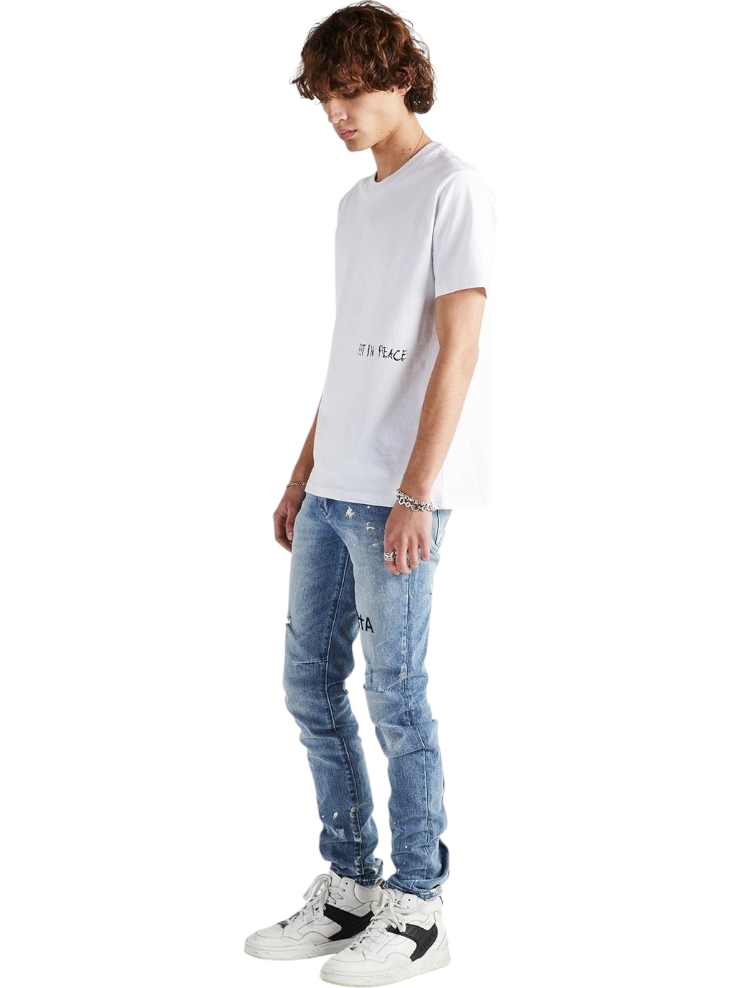 A person stands against a white background wearing an RTA Clayton Jean Distressed White Paint Blue, a white t-shirt, and white sneakers, looking downward.