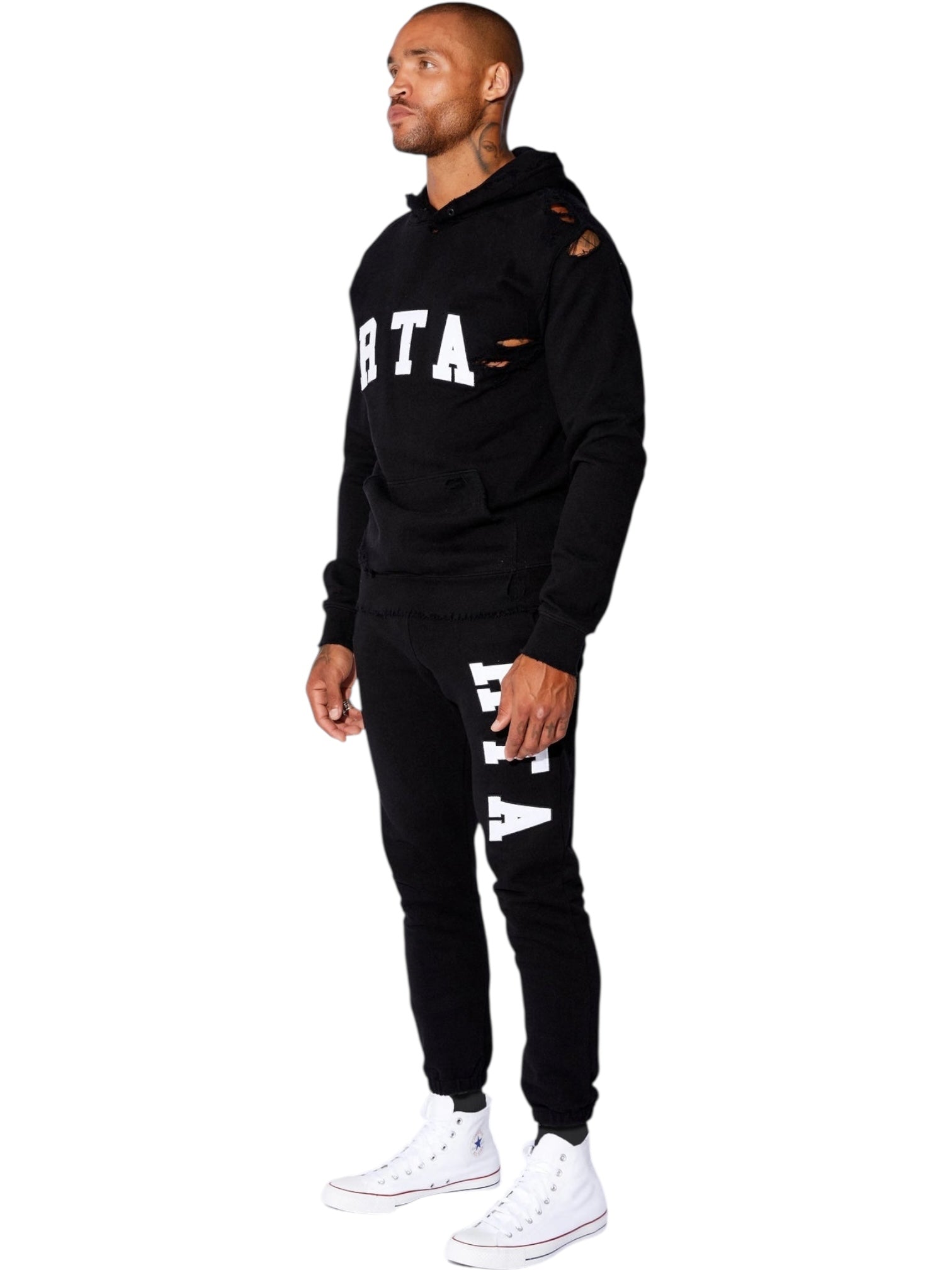 A man stands against a white background wearing RTA Owen black collegiate sweatpants and a matching hoodie, both with white RTA logos, paired with white high-top sneakers.