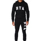 A man stands facing forward in the RTA Owen black collegiate hoodie and sweatpants with white RTA logos, paired with white high-top sneakers, against a white background.