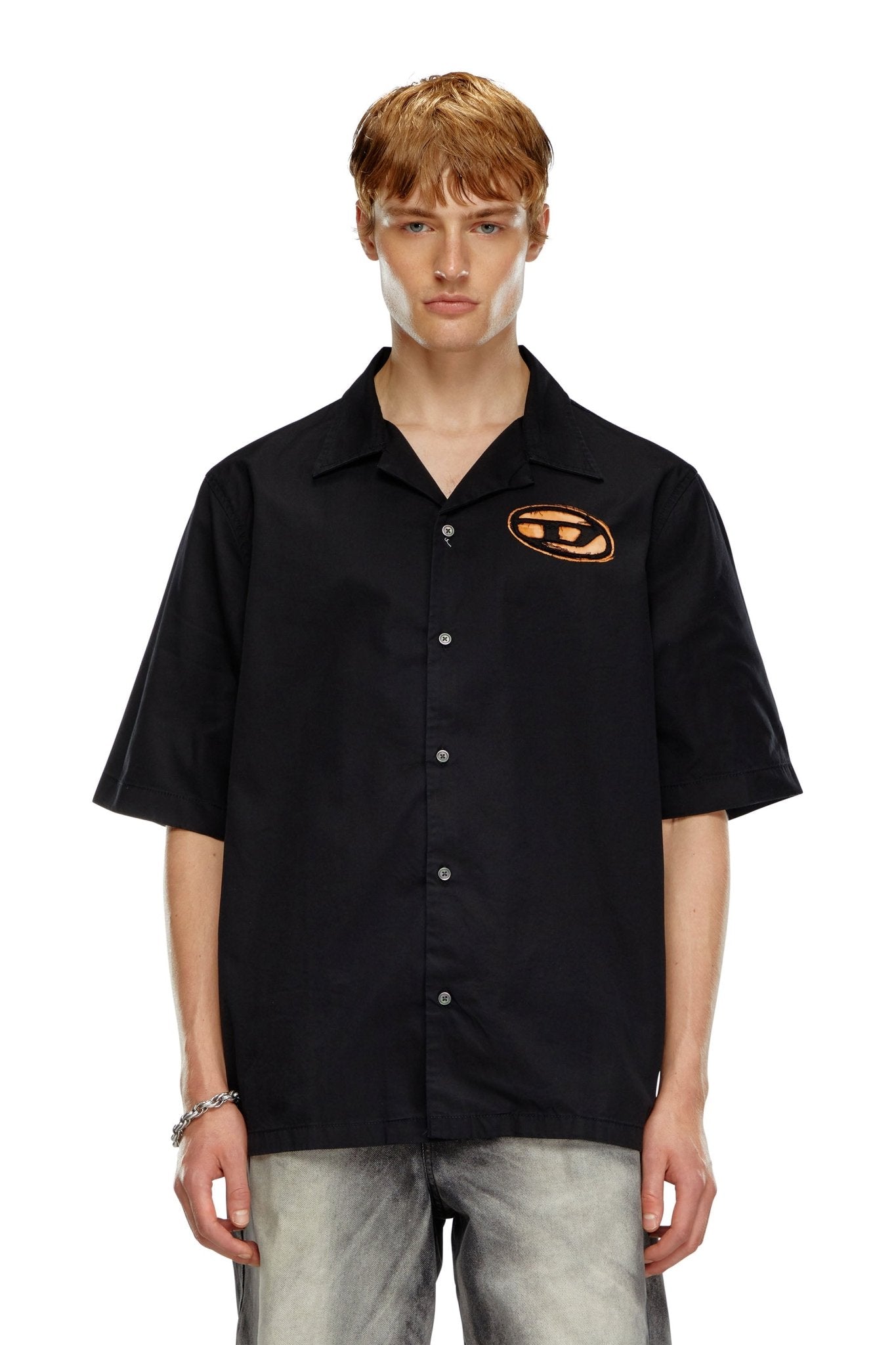 A person with short blonde hair is wearing the stylish Diesel S-Stan Bleach Black bowling shirt by DIESEL, which features a modern orange design on the chest, paired with light gray pants. The sharp camp collar of the shirt enhances its contemporary look against the plain white background.