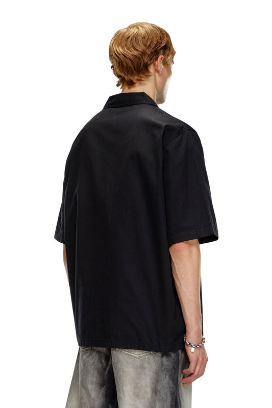 A person with light hair is wearing the Diesel S-Stan Bleach Black short-sleeved bowling shirt made from organic cotton, paired with gray pants, and is facing away from the camera.