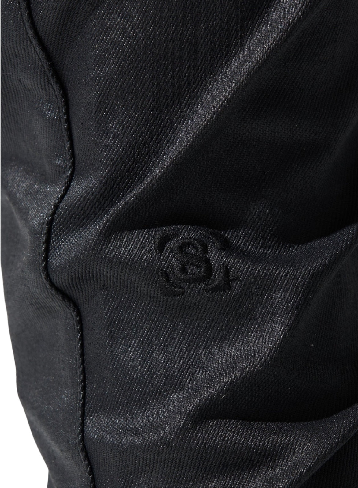 Close-up of black wax wash fabric with a stitched symbol featuring a circle and a stylized "S." The material appears to be slightly textured, reminiscent of SERENEDE CAVIAR WAX STACKED JEANS BLK by SERENEDE, and has subtle creases.