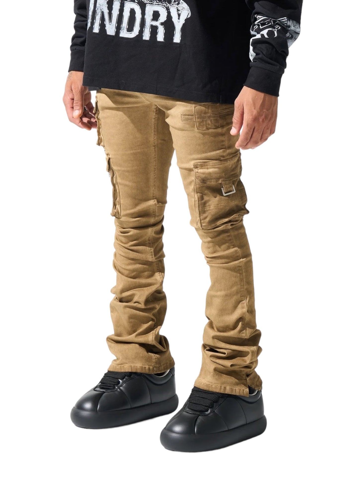 Person wearing SERENEDE CINNAMON CARGO STACKED BRWN pants with a leather logo patch, black sneakers, and a black long-sleeve shirt with white text. The pants have multiple pockets and appear slightly oversized at the ankles, reminiscent of the SERENEDE stacked cargo jeans style.