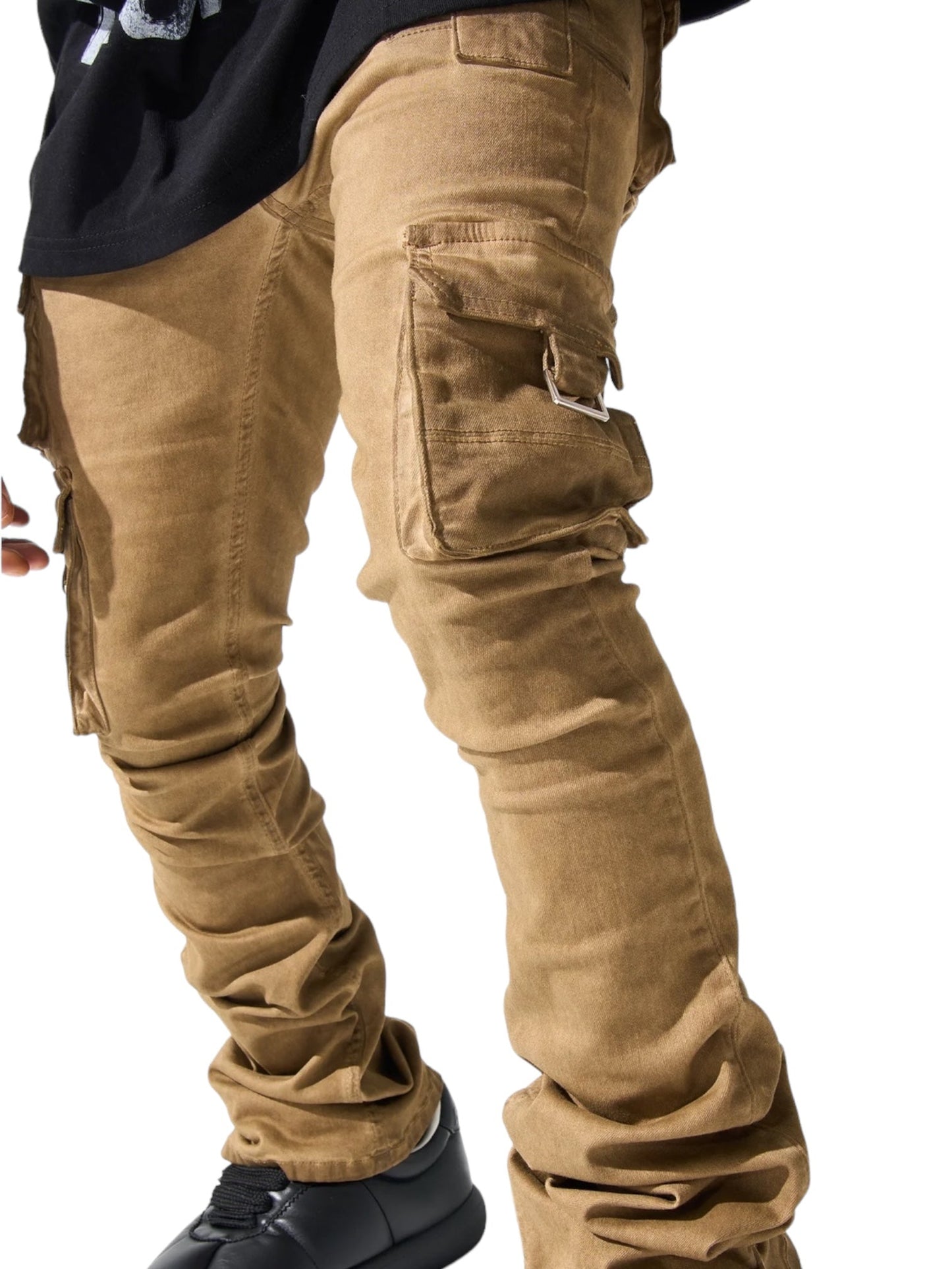 Close-up of a person wearing SERENEDE CINNAMON CARGO STACKED BRWN jeans and black shoes, with legs slightly apart. The jeans feature multiple pockets, including a flap pocket on the thigh adorned with a leather logo patch. The background is plain white.