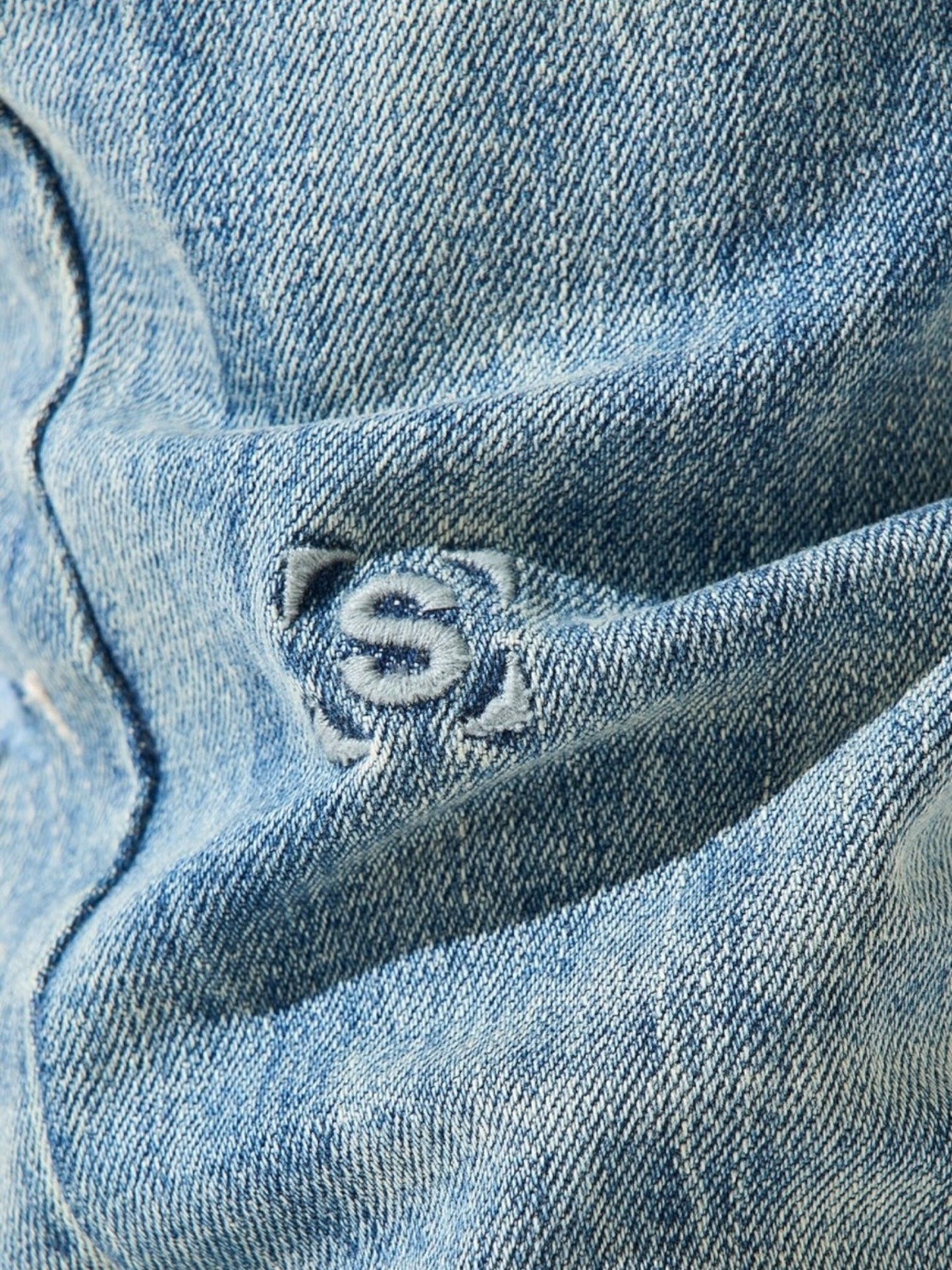 Close-up of the Serenede Imperial Straight Leg Jeans Blue by SERENEDE, highlighting the classic blue denim fabric adorned with an embroidered dollar sign.